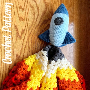 May include: A crocheted rocket toy in light blue with dark blue fins and a gray porthole sits atop a colorful crocheted blanket. The blanket features a flame pattern in orange, yellow, white, and gray. The words "Crochet Pattern" are written in white.