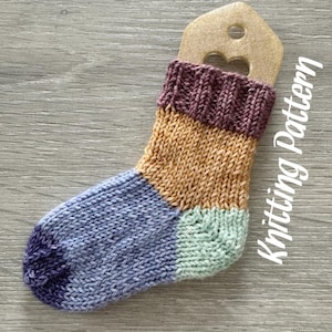 May include: A colorful knitted sock with a purple, orange, and green color scheme. The sock is hanging on a wooden tag with the text "Knitting Pattern".