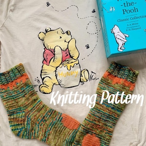 May include: Knitting pattern for a pair of socks in a green, orange and blue colour scheme. The socks are laid out on a beige t-shirt with a Winnie the Pooh graphic. The t-shirt has the text "Winnie-the-Pooh Classic Collection" on it.