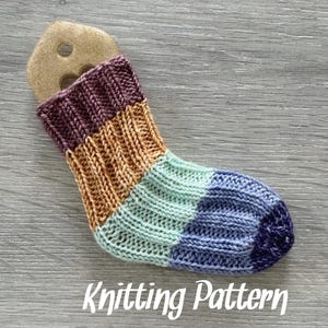 Puede incluir: Un calcetín de punto con un patrón de rayas en tonos de morado, naranja, verde y azul. El calcetín está sobre una superficie de madera con el texto "Knitting Pattern" debajo.
