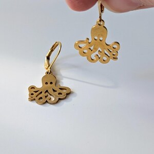 Könnte beinhalten: Ein Paar goldfarbene Ohrringe mit einem Kraken-Design. Die Ohrringe sind aus Metall und haben einen baumelnden Stil.