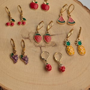Könnte beinhalten: Ein Set mit 10 goldfarbenen, baumelnden Ohrringen in Form von Früchten. Die Ohrringe zeigen eine Vielzahl von Früchten, darunter Äpfel, Kirschen, Wassermelonen, Erdbeeren, Ananas und Trauben. Die Ohrringe sind alle aus Emaille gefertigt und haben eine glänzende Oberfläche.