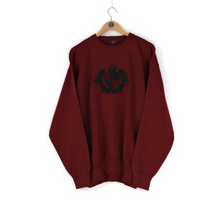 May include: A burgundy crew neck sweater with a black embroidered crest design. The sweater is hanging on a wooden hanger against a white background. The crest features a crown and shield.