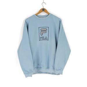 Puede incluir: Una sudadera azul claro con un logotipo FILA blanco en un cuadro negro.
