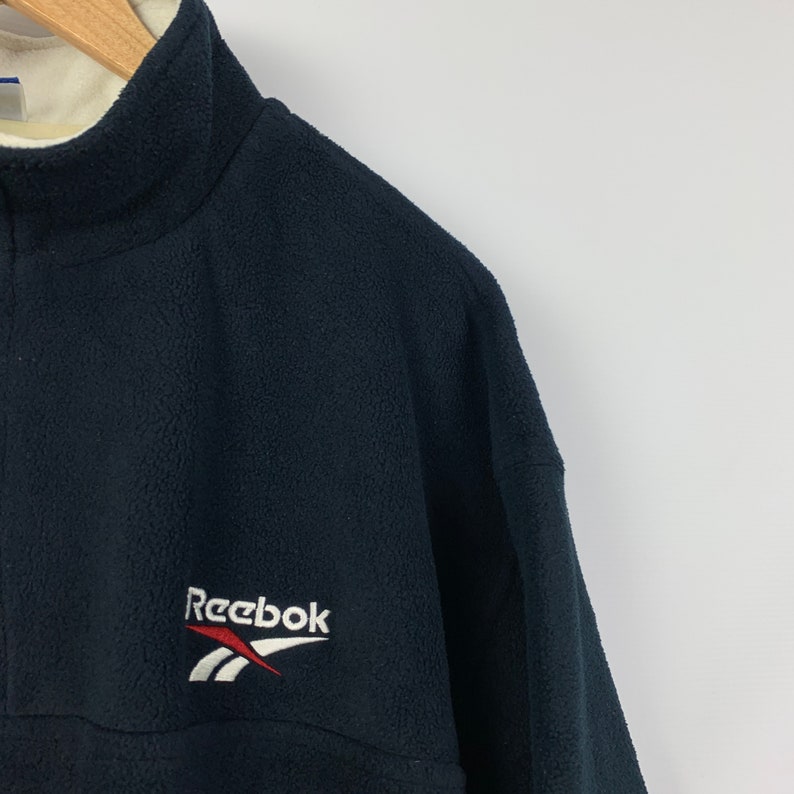 May include: Black fleece pullover with a white Reebok logo and red accents. The zipper is partially visible.