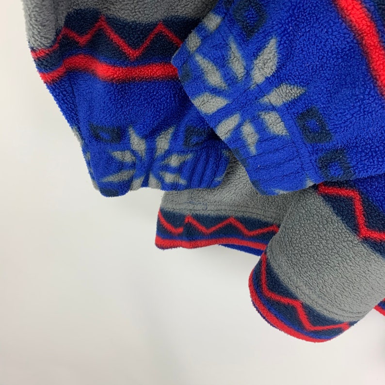 Puede incluir: Primer plano de una chaqueta de forro polar azul y gris con un patr&oacute;n geom&eacute;trico. La chaqueta tiene un ribete rojo y azul.