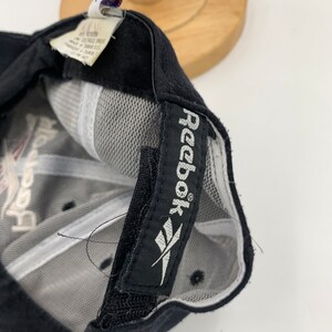 May include: Black baseball cap with a white Reebok logo on the back. The cap has a mesh back and a black adjustable strap.