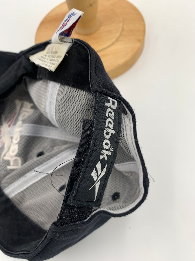 May include: Black baseball cap with a white Reebok logo on the back. The cap has a mesh back and a black adjustable strap.