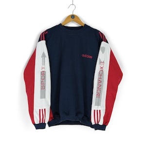 Pode incluir: Camisola Adidas azul marinho com detalhes vermelhos nas mangas e ombros. As mangas apresentam painéis brancos com as palavras "SPORT" e "EXCHANGE" em vermelho. O logótipo Adidas é vermelho.