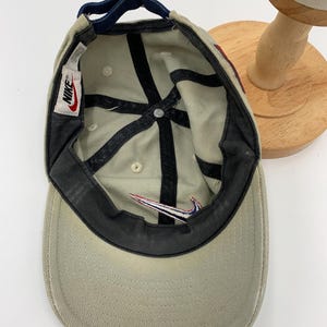 Może przedstawiać: Beżowa czapka z daszkiem z regulowanym granatowym paskiem. Czapka ma czarny wewnętrzny rondo i biało-czerwone logo Nike. Wnętrze ma białą metkę Nike.