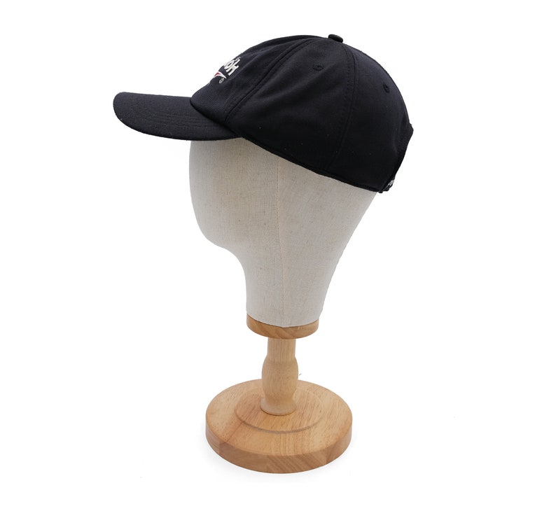 May include: A black baseball cap with a white logo on the front. The cap is on a wooden display stand.
