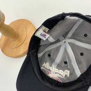 May include: Black mesh baseball cap with a white Reebok logo embroidered on the front. The cap has a black adjustable strap.