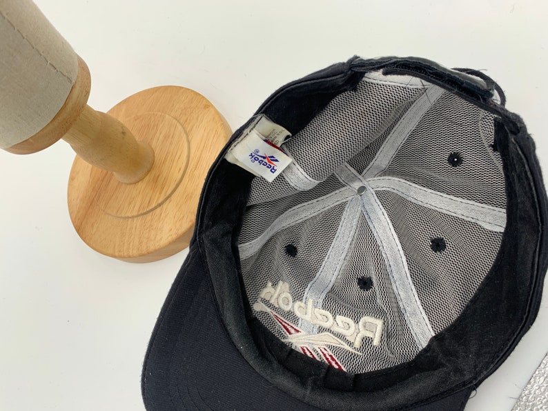May include: Black mesh baseball cap with a white Reebok logo embroidered on the front. The cap has a black adjustable strap.