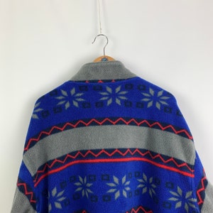 Puede incluir: Una chaqueta de forro polar azul y gris con un patr&oacute;n geom&eacute;trico. El patr&oacute;n presenta copos de nieve, diamantes y zigzags en rojo, azul y gris.