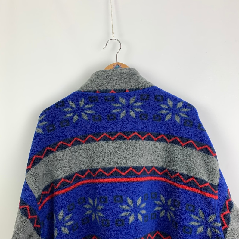 Puede incluir: Una chaqueta de forro polar azul y gris con un patr&oacute;n geom&eacute;trico. El patr&oacute;n presenta copos de nieve, diamantes y zigzags en rojo, azul y gris.