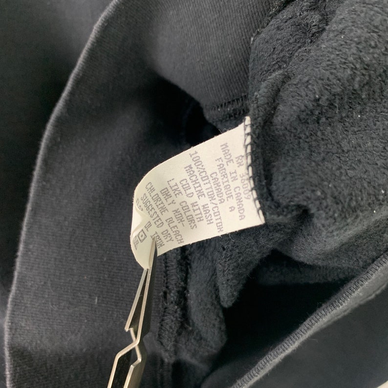 May include: A black care tag with white text that reads: "RN 36009 Made in Canada Fabrique a Canada 100% Cotton/Coton Machine Wash Cold With Like Colors Only Non-Chlorine Bleach Suggested Dry"