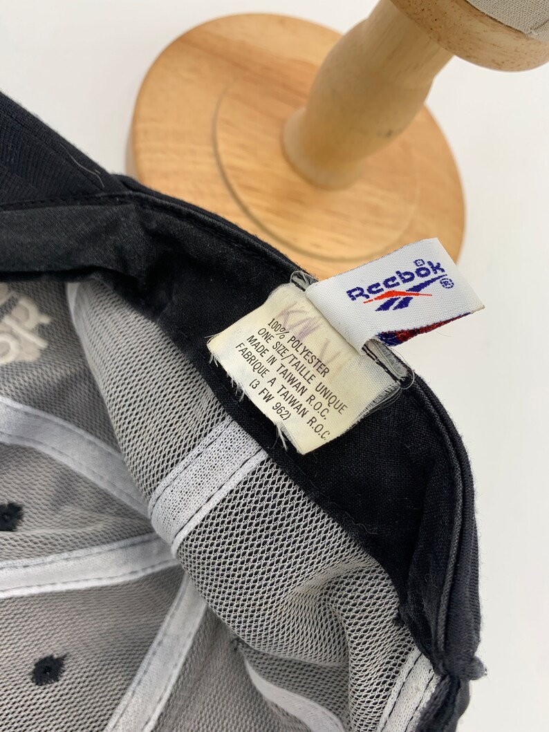 May include: Black mesh baseball cap with a white Reebok logo on the front and a white tag on the back that reads "100% Polyester, One Size/Taille Unique, Made in Taiwan R.O.C., Fabrique a Taiwan R.O.C., (3 FW 962)"