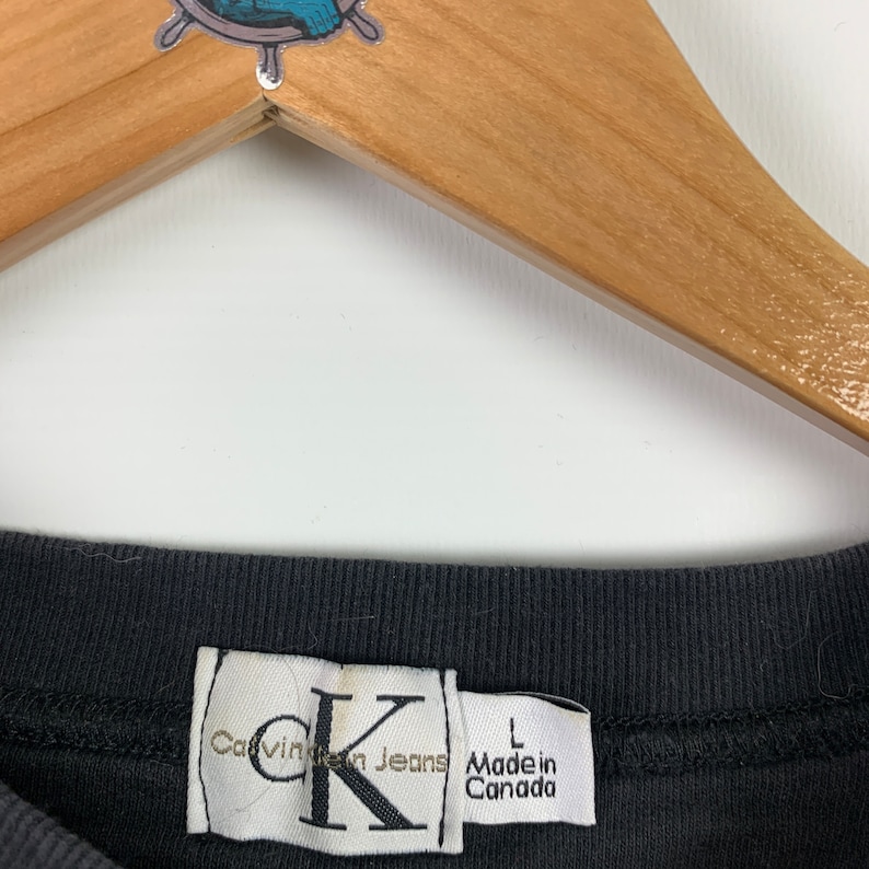 May include: Black t-shirt with a white Calvin Klein Jeans tag. The tag features a large "K" and the text "Calvin Klein Jeans" and "Made in Canada" in black text. The tag also has a size "L" printed on it.