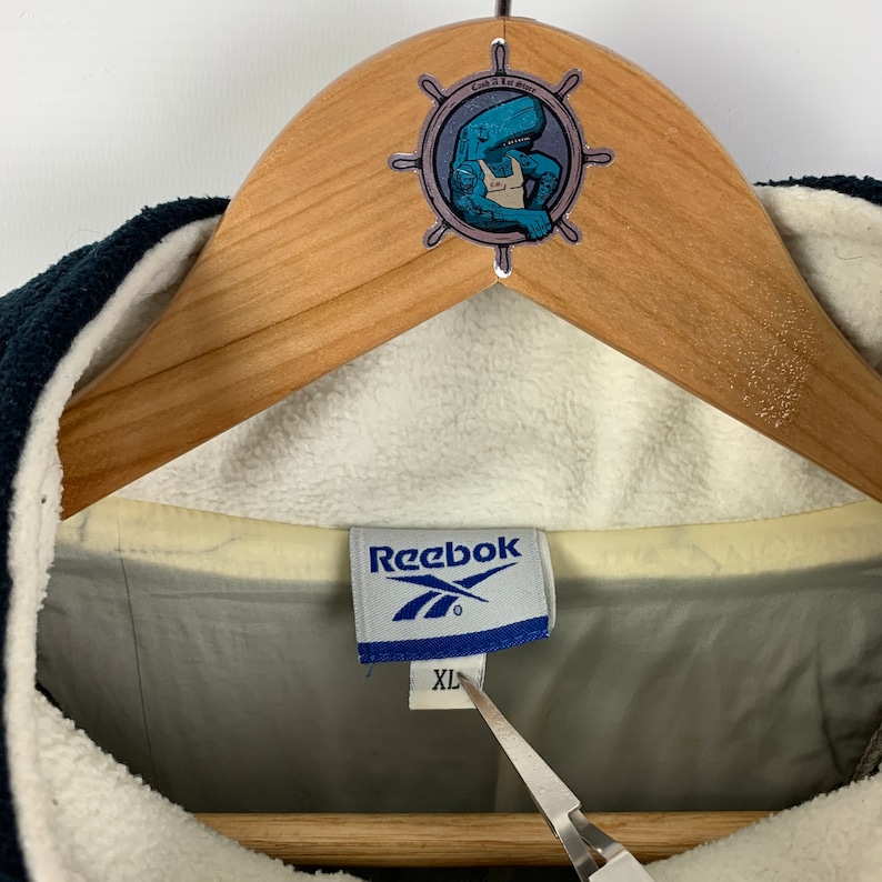 May include: A blue Reebok brand label with white text and a white logo. The label is attached to a white and gray jacket with a black zipper. The label says "Reebok" and "XL".