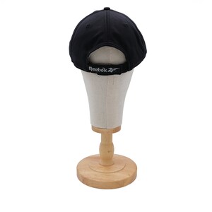 May include: Black baseball cap with a white Reebok logo on the back. The cap is displayed on a wooden mannequin head.
