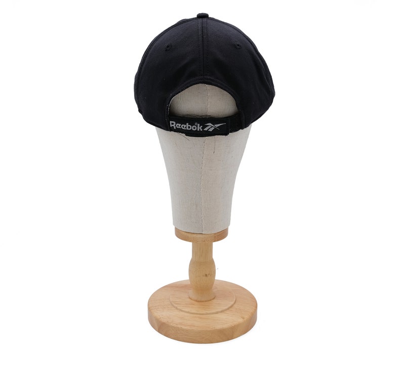 May include: Black baseball cap with a white Reebok logo on the back. The cap is displayed on a wooden mannequin head.