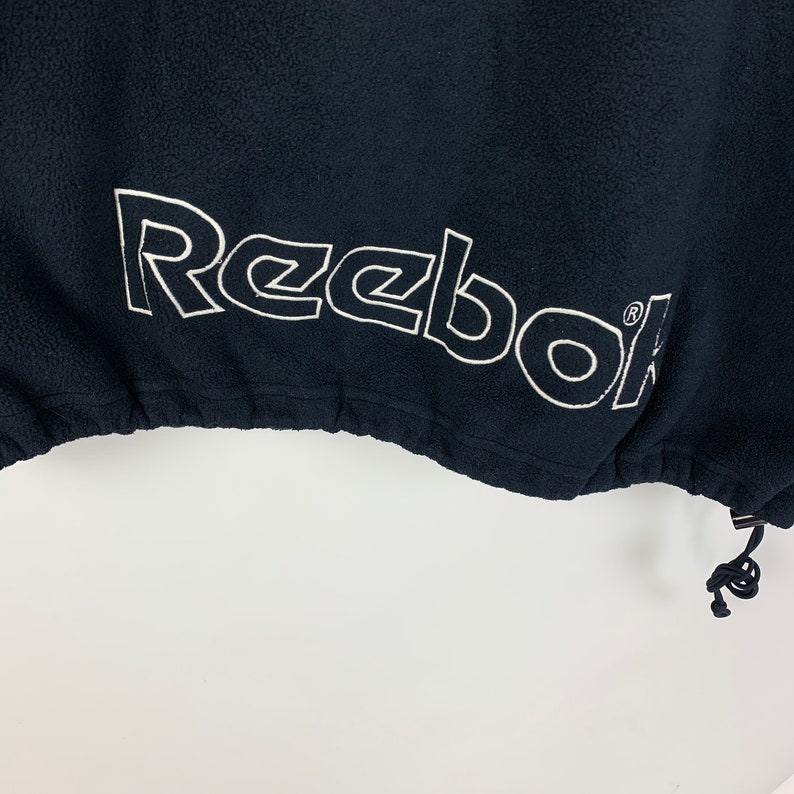 May include: Black fleece jacket with a white Reebok logo embroidered on the bottom hem. The jacket has a drawstring at the bottom.