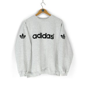 Puede incluir: Una sudadera Adidas gris claro con letras y logotipos negros. La palabra "adidas" está centrada en el pecho. El logotipo del trébol Adidas está en las mangas izquierda y derecha. La sudadera está colgada en una percha de madera.
