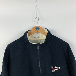 May include: A black fleece pullover with a half-zip closure and a white Reebok logo on the front.