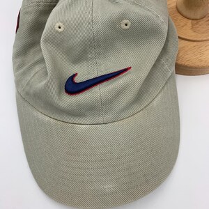 Może przedstawiać: Jasnobrązowa czapka z daszkiem z zakrzywionym daszkiem. Czapka posiada granatowe i czerwone haftowane logo Nike swoosh z przodu. Czapka ma guzik na g&oacute;rze i otwory wentylacyjne.