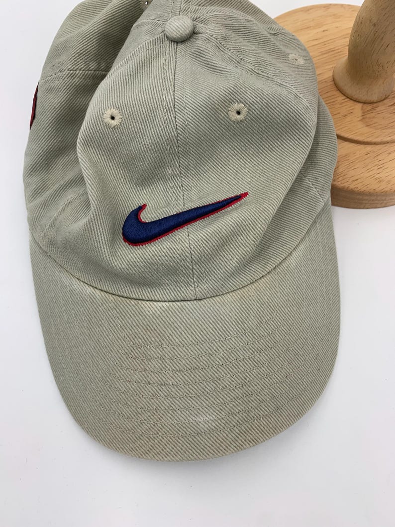 Może przedstawiać: Jasnobrązowa czapka z daszkiem z zakrzywionym daszkiem. Czapka posiada granatowe i czerwone haftowane logo Nike swoosh z przodu. Czapka ma guzik na g&oacute;rze i otwory wentylacyjne.