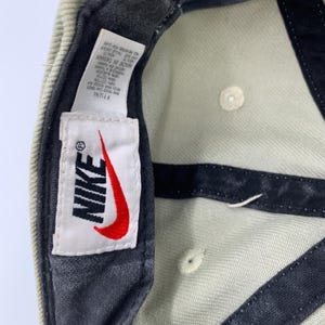 Może przedstawiać: Jasnozielona czapka z daszkiem Nike z czerwonym logo Swoosh na białej etykiecie. Czapka ma ciemnoszary daszek i niebieski pasek. Na etykiecie widnieje napis "NIKE" i "MADE IN TAIWAN".