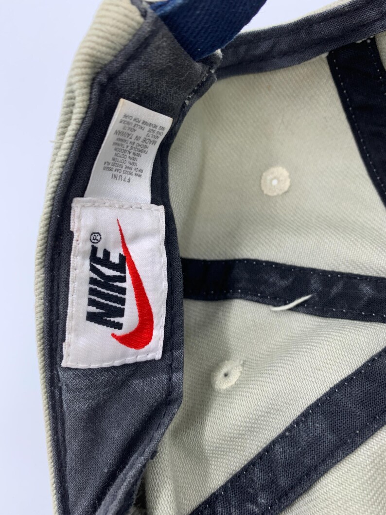 Może przedstawiać: Jasnozielona czapka z daszkiem Nike z czerwonym logo Swoosh na białej etykiecie. Czapka ma ciemnoszary daszek i niebieski pasek. Na etykiecie widnieje napis "NIKE" i "MADE IN TAIWAN".