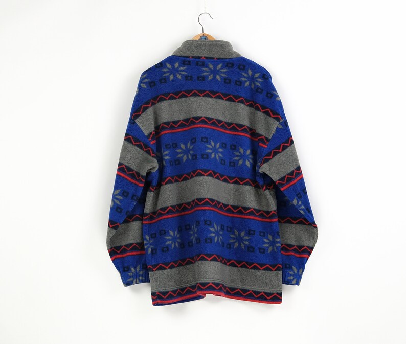 Puede incluir: Una chaqueta de forro polar azul y gris con un patr&oacute;n geom&eacute;trico de copos de nieve. La chaqueta tiene una cremallera y mangas largas.