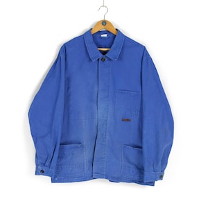 Op de afbeelding: Blauw werkjack met een knoopsluiting, twee voorzakken en twee lagere zakken. De jas hangt aan een houten hanger. De jas heeft een klein rood label op de rechterzak.