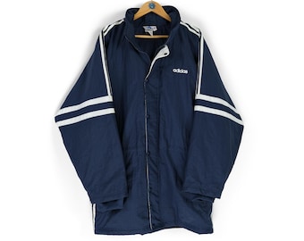 May include: Navy blue Adidas jacket with white stripes on the sleeves and shoulders. The jacket has a button-up front and a high collar. The Adidas logo is visible on the chest. The jacket is hanging on a wooden hanger.