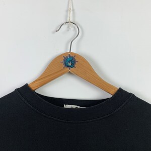 May include: Black crew neck sweatshirt hanging on a wooden hanger with a nautical themed sticker. The sticker features a blue and white illustration of a sailor with a ship's wheel.
