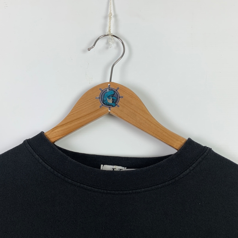 May include: Black crew neck sweatshirt hanging on a wooden hanger with a nautical themed sticker. The sticker features a blue and white illustration of a sailor with a ship's wheel.