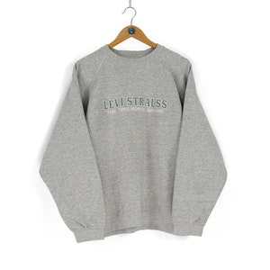Pode incluir: Um moletom cinza Levi Strauss com o texto "LEVI STRAUSS THE TWO HORSE BRAND" em azul claro. O moletom está pendurado em um cabide de madeira em um fundo branco. O moletom tem mangas compridas e gola redonda.