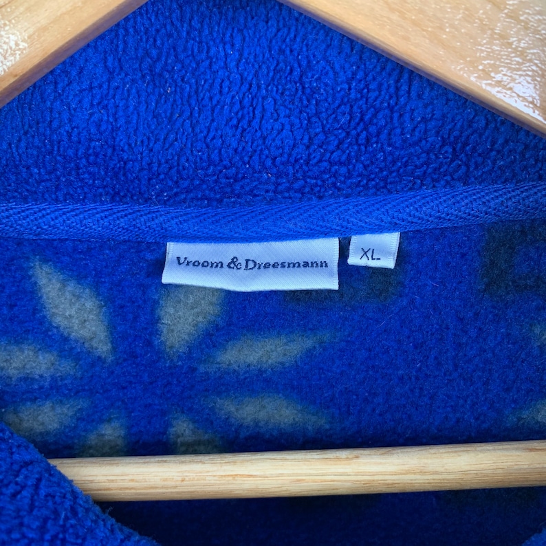 Puede incluir: Una chaqueta de forro polar azul con una etiqueta blanca que dice "Vroom & Dreesmann" y "XL". La chaqueta tiene un estampado floral.