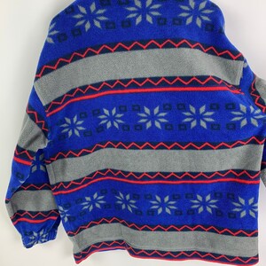 Puede incluir: Chaqueta de forro polar azul y gris con un patr&oacute;n de copo de nieve. La chaqueta tiene una cremallera y un cuello de pie.