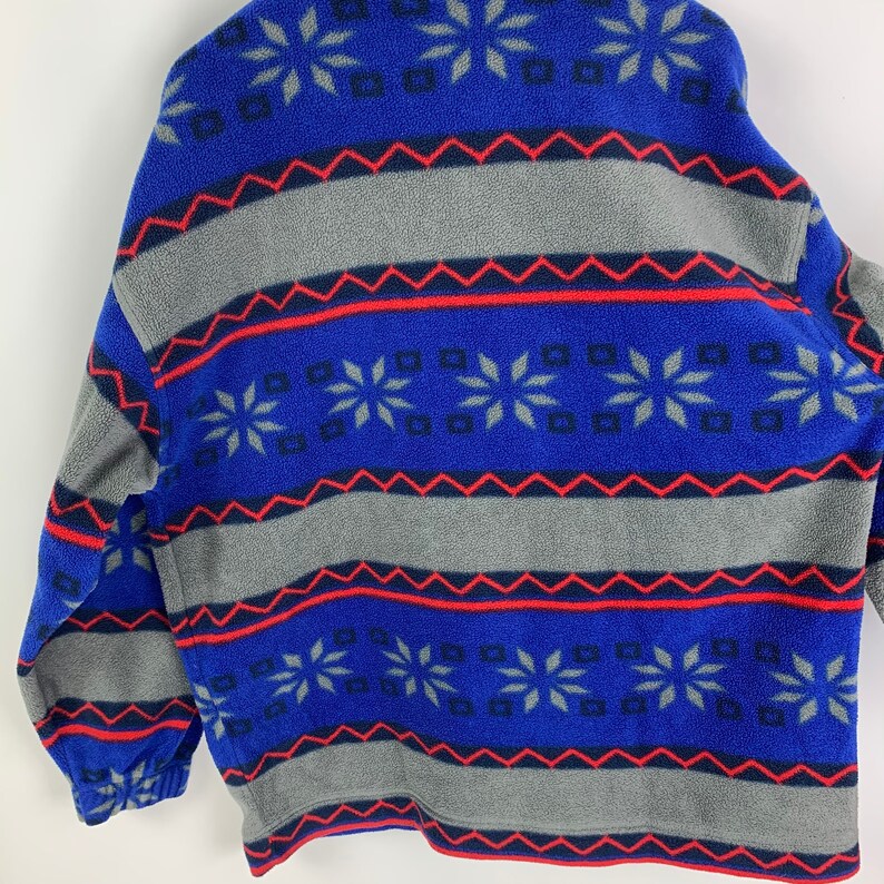 Puede incluir: Chaqueta de forro polar azul y gris con un patr&oacute;n de copo de nieve. La chaqueta tiene una cremallera y un cuello de pie.