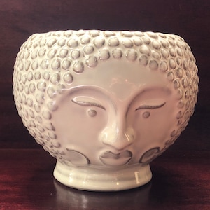 May include: A white ceramic planter shaped like a face with a textured surface. The face has closed eyes, a nose, and a slightly parted mouth.