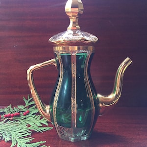 May include: A green glass teapot with gold accents and a gold lid. The teapot has a round body and a spout on the side. The teapot is sitting on a wooden surface.