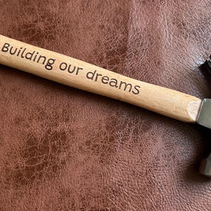 Puede incluir: Un martillo con mango de madera y cabeza de metal plateado. El mango está grabado con las palabras "Building our dreams".