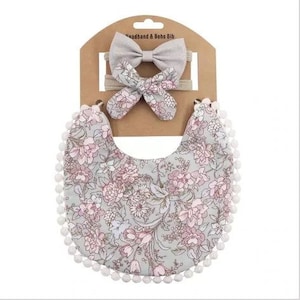 Op de afbeelding: Een set van twee grijze stoffen hoofdbanden met bloemenprint en een slabbetje met bloemenprint en witte pomponrand. De verpakking bevat de tekst "Headband & Boho Bib".