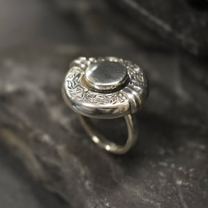 May include: A silver ring with a round, engraved top and a smooth, circular centre. The ring is set on a dark, textured surface.