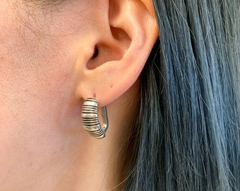 May include: Silver hoop earrings with a textured, striped design. The earrings are worn in a person's ear.