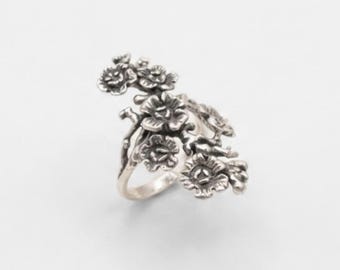 Puede incluir: Un anillo de plata con un diseño detallado y tridimensional de rama floral. El metal tiene un acabado oxidado que resalta las texturas de las múltiples flores pequeñas y tallos que envuelven el aro y se extienden hacia el exterior.