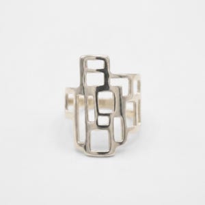 May include: A silver-colored ring with a geometric design. The ring features a series of rectangular and square cutouts, creating an open, abstract pattern. The ring is set against a white background, highlighting its modern design.