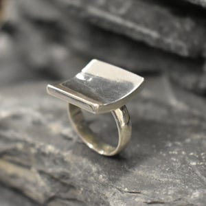 Puede incluir: Un anillo de plata con una banda ancha, plana y rectangular. La banda está curvada en la parte superior e inferior, lo que le da una forma única.